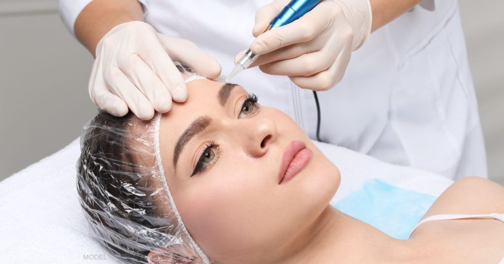 A girl with her hair in a clear cap is laying down while someone rests their hands her face as she is starting to microblade the eyebrows of the patient. (model)